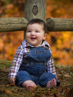 Tuscaloosa photographer picture of a baby in overalls against a fence with beautiful fall colors.