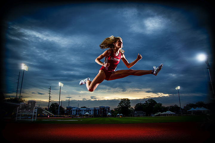 University of Alabama Graduation Pictures of a track and field athlete in her Alabama Track uniform leaping it the track lights at dusk.